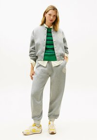 Mujer con chaqueta gris abotonada y pantalones deportivos a juego sobre suéter verde y camisa a rayas, combinados con zapatillas amarillas y blancas.