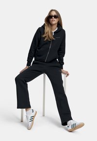 Svart zip-up hettegenser med frontlommer, sammen med matchende joggebukser. Modellen har på seg hvite sneakers med grønne striper og solbriller.