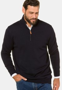 Maglione navy a maglia con zip a quarto, caratterizzato da una texture a costine, colletto rialzato e un girovita in pelle sulla cerniera. Abbinato a una camicia bianca sotto.