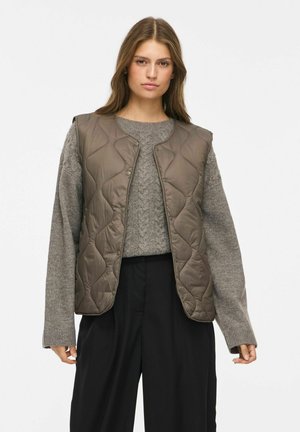 Kvinde med langt brunt hår iført en grå strikket sweater, olivengrøn quiltet vest og løse sorte bukser mod en ensfarvet baggrund.