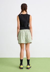 Top negro sin mangas, pantalones cortos cargo verde claro con cintura elástica y calcetines negros con zapatillas amarillas y rojas. Fondo liso.