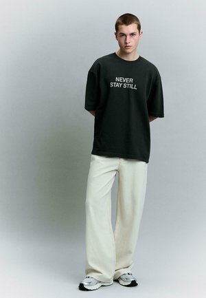 Jongere staat met handen achter de rug, draagt een zwart oversized T-shirt met de tekst "NEVER STAY STILL", wijde witte broek en witte sneakers.