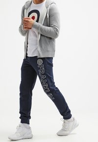 Grå zip-up hoodie över en vit t-shirt med ett cirkulärt mönster, tillsammans med marinblå sweatpants med "FRANKLIN" grafik, och vita sneakers.