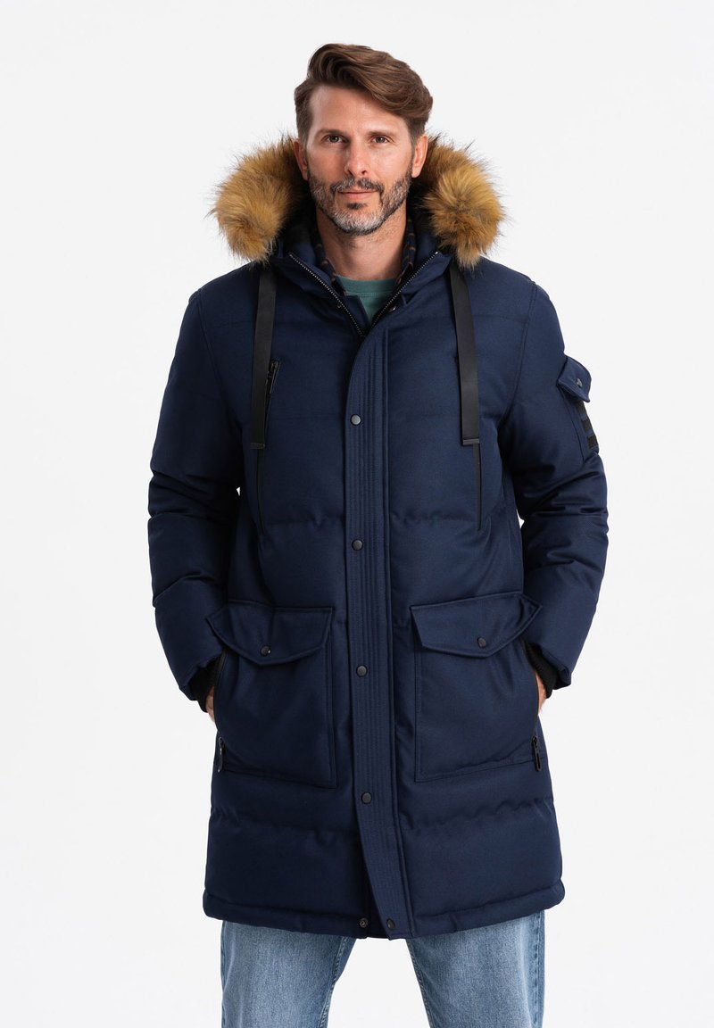 Parka imbottita blu navy con cappuccio rifinito in pelliccia, cinghie nere e numerose tasche; una superficie liscia e un design fino al ginocchio.