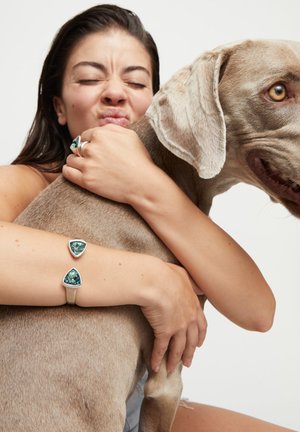 Femme aux cheveux foncés embrassant un grand chien brun clair, portant des bijoux en argent avec des pierres précieuses vertes sur le doigt et le poignet.