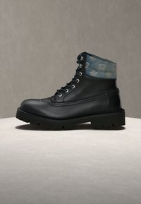Bottine en cuir noir avec une semelle épaisse et des lacets noirs, avec un revers en denim bleu présentant un motif blanc délavé autour du haut.