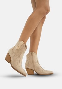 Botas de tobillo beige de ante con adornos decorativos, puntera afilada y tacón grueso. Cremallera lateral y detalle de costura de estilo western.