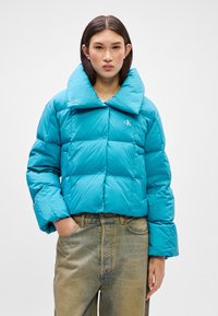 Tirkizna puffer jakna s velikim ovratnikom, kockastim uzorkom i logotipom na prsima. Uparen s hlačama od svijetlog denima visokog struka.