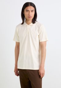 Polo a maniche corte di colore crema realizzato in tessuto morbido, con colletto classico e due bottoni, decorato con un discreto logo ricamato sul petto.