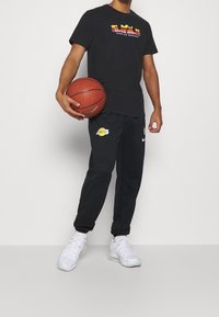 Camiseta negra de manga corta con el colorido texto "DEFENDER LA GRANDEZA", combinada con pantalones de chándal negros que tienen el logo de los Lakers, zapatillas blancas y sosteniendo un balón de baloncesto.