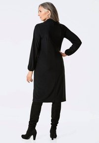 Schwarzes langärmliges Kleid mit gerader Silhouette, subtilen Drapierungen und einem Reißverschluss im Rücken. Kombiniert mit kniehohen schwarzen Wildlederstiefeln.