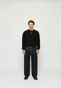 Černé tričko s dlouhým rukávem spojené s tmavými džínami a černými botami, s jednoduchým designem a neutrálními barvami, stojící před bílým pozadím.