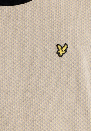 Nærbillede af beige tekstureret stof med et lille gentaget diamantmønster og en gul broderet fuglelogo på brystområdet.