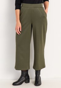 Olivgrüne, weit geschnittene Hose aus weichem Stoff, mit seitlichen Taschen und einem glatten Bündchen, kombiniert mit schwarzen Stiefeletten.