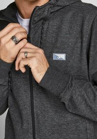 Sudadera con cremallera gris de acabado texturizado, con una etiqueta azul y cordones en contraste. Manos con anillos parcialmente visibles.