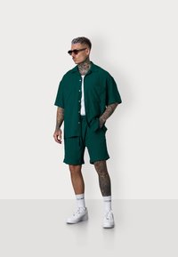 Man stands in a green short-sleeve shirt and matching shorts, wearing sunglasses, white tank top, and sneakers, with tattoos visible on his arms.