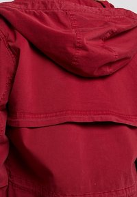 Giacca rossa con cappuccio caratterizzata da una vestibilità rilassata, dettagli cuciti e un design a strati nella parte posteriore con una texture liscia.