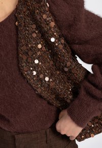 Persoon in een fuzzy donkerbruin trui die een gestructureerde, met pailletten versierde clutchtas in bruin en meerdere kleuren tegen hun zij houdt.