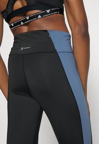 Leggings neri con un pannello laterale blu, dotati di una tasca con zip e branding "AEROREADY". Il tessuto è liscio e presenta un design aderente.