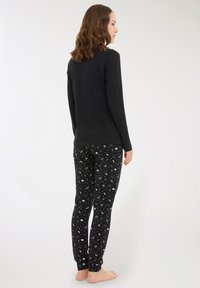Donna in piedi a piedi nudi, con il corpo di lato, indossa un top lungo nero e pantaloni da pigiama neri con motivi celestiali bianchi.