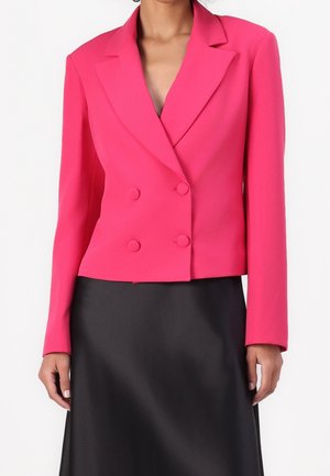 Femme portant un blazer rose vif à double boutonnage avec des revers crantés, sur une jupe noire, debout devant un fond blanc.