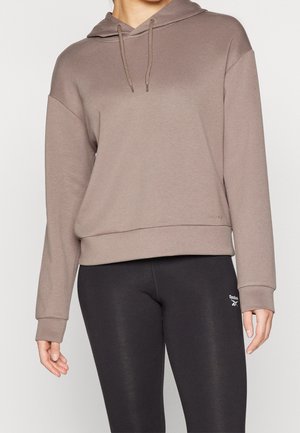 Personne portant un sweat à capuche couleur taupe et des leggings noirs Reebok avec le logo de la marque visible sur la cuisse gauche.