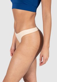 Sous-vêtements en tanga beige avec un design sans coutures, texture douce et taille basse. Présente une finition en tissu lisse et une couverture minimale.