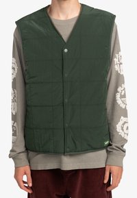 Gevoerde groene vest met een V-hals, drukknoopjes en een lichtgewicht, gestructureerde stof. Draag het over een grijs shirt met lange mouwen en witte graphics.