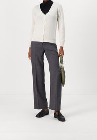 Osoba v bílé zapínací kardiganu přes černý rolák, šedých širokých kalhotách, černých mokasínech, držící olivově zelenou kabelku na boku.