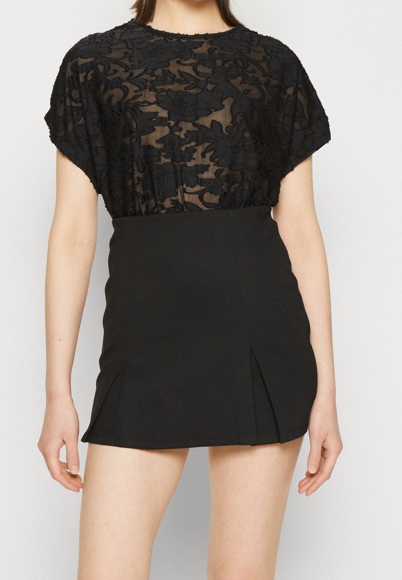 Top negro de encaje con patrón floral y mangas cortas, superpuesto sobre una mini falda negra ajustada con plisados frontales y una textura suave.