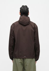 Άτομο με πλεξούδες που φοράει σκούρο καφέ μπουφάν με κουκούλα και χακί παντελόνι, στέκεται γυρισμένο με την πλάτη προς το μέρος ενός απλού φόντου.