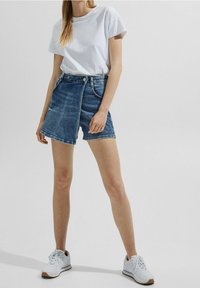 Jeansshorts in hellem Blau mit hoher Taille, vorderer Naht und lockerem Schnitt. Kombiniert mit einem schlichten weißen T-Shirt und weißen Turnschuhen.
