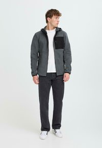 Mann in grauer Zip-Jacke aus Fleece mit schwarzer Brusttasche, weißem T-Shirt, dunkler Jeans und weißen Sneakers, der seitlich schaut.