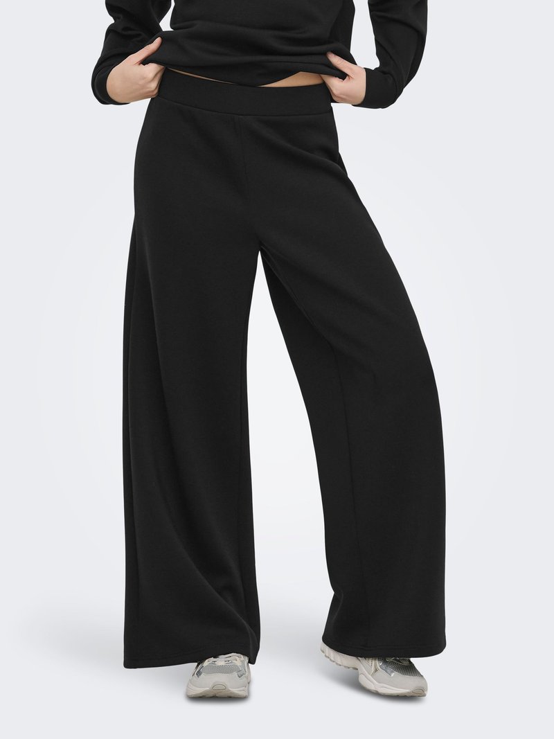 Persoon draagt zwarte wijde broekspijpen, zwarte top die iets opgetild is, en grijze sneakers, staand tegen een eenvoudige lichte achtergrond.