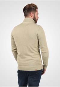 Beige gebreide trui met een hoge kraag, geribde manchetten en een onderrand. Heeft een subtiele textuur en een klein logopatch aan de achterkant.