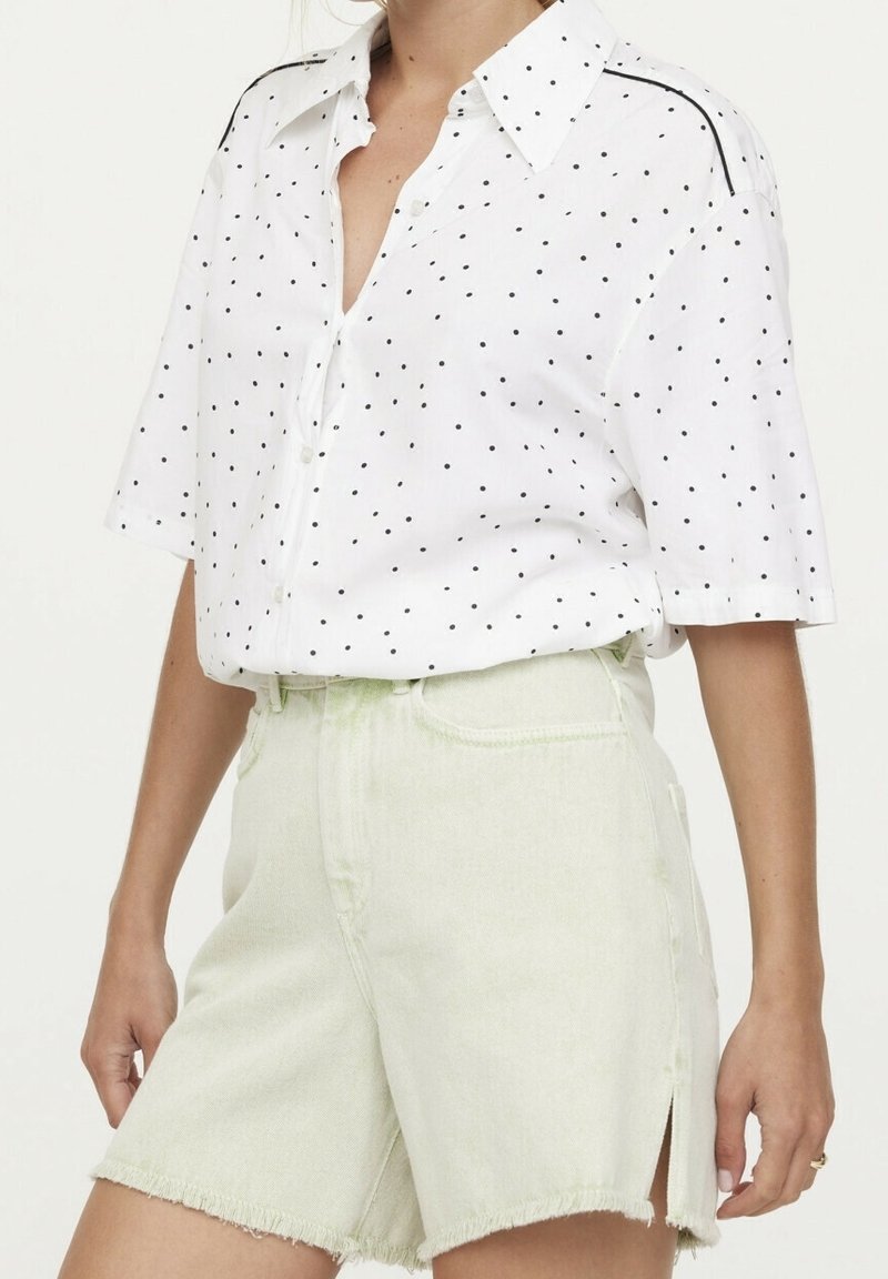Chemise blanche à boutons avec des pois noirs, manches courtes et détails contrastants aux épaules, associée à un short en jean vert clair.