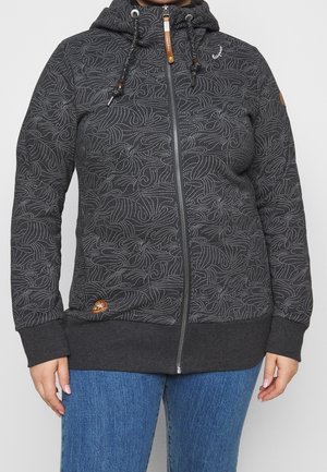 Woman wearing a dark gray zip-up hoodie with white abstract dotted patterns and blue jeans against a plain background.