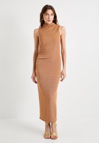 Kampas, gaun bodycon tanpa lengan dengan leher draping, tekstur halus, dan siluet panjang, dipadukan dengan sandal bertali emas.