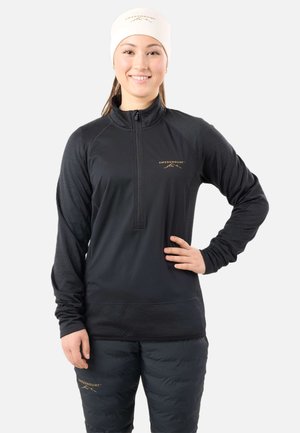 Jeune femme portant une veste et un pantalon de plein air noirs Swedemount avec un bonnet blanc, souriant, une main sur la hanche, sur fond blanc.