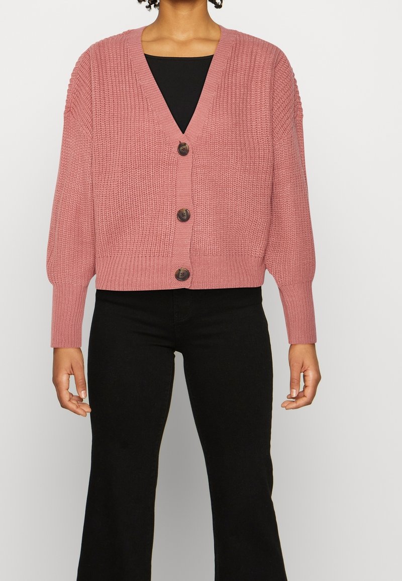 Femme portant un cardigan épais tricoté rose poudré avec trois boutons, par-dessus un haut noir et un pantalon noir, debout devant un fond uni.
