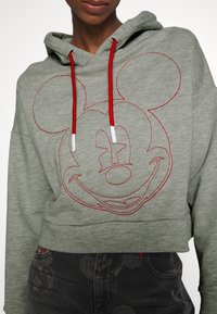 Personne portant un sweat à capuche court gris clair avec le visage de Mickey Mouse brodé en rouge et des cordons rouges, associée à un pantalon noir avec des imprimés subtils de Mickey.
