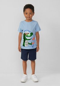 Hellblaues Baumwoll-T-Shirt mit einem grünen Krokodil-Graphic, das mit Pailletten verziert ist, kombiniert mit marineblauen Shorts und weißen Sneakers.
