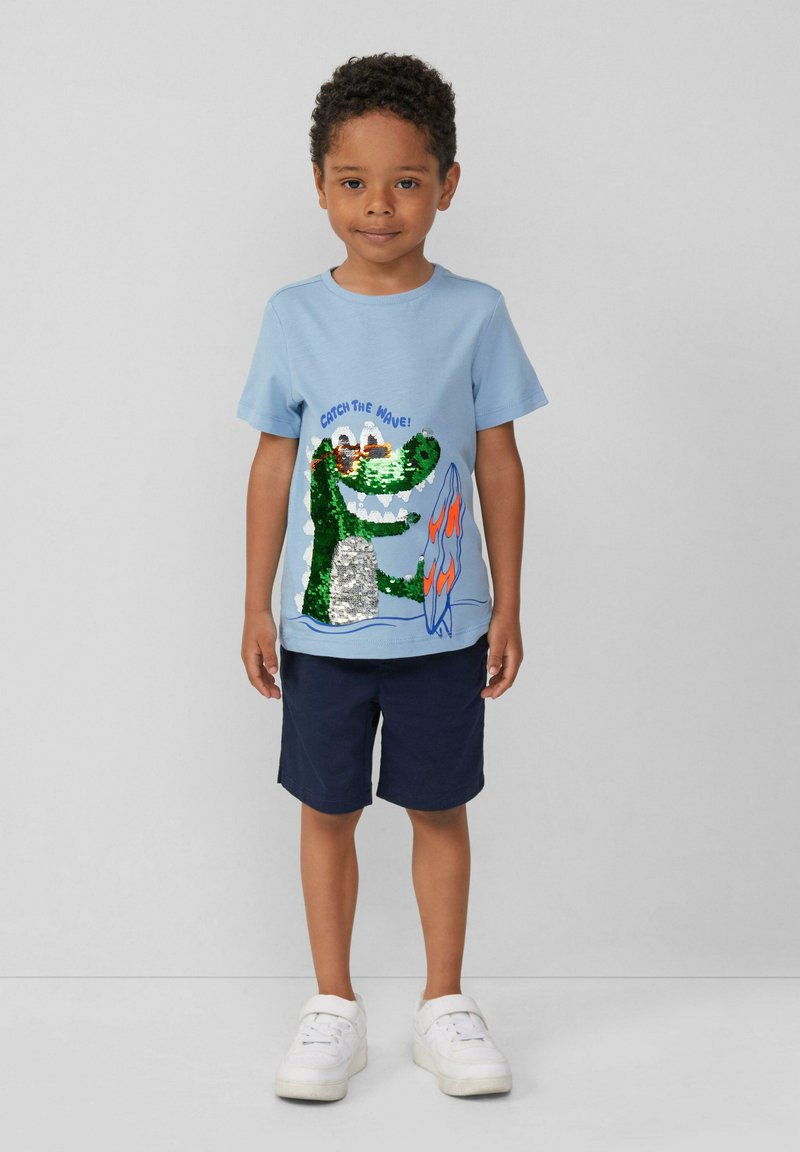 Hellblaues Baumwoll-T-Shirt mit einem grünen Krokodil-Graphic, das mit Pailletten verziert ist, kombiniert mit marineblauen Shorts und weißen Sneakers.