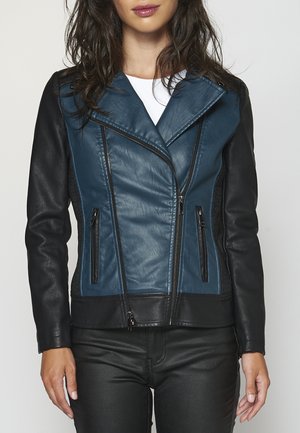 Femme portant une veste en cuir bleue et noire par-dessus un haut blanc, debout les bras détendus le long du corps devant un fond uni.