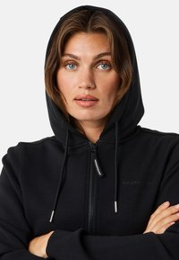 Zwarte hoodie met een ritssluiting aan de voorkant, verstelbare koordkap en een zachte textuur. Voorzien van een subtiele logo op de borst en metalen accenten.
