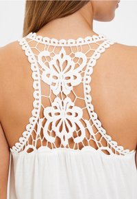 Bílý top s detailním krajkovým zadním dílem s květinovými vzory, vlnitými okraji a volným tělem z měkké tkaniny.
