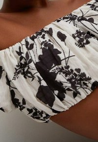 Haut décolleté épaules en tissu blanc léger orné de motifs floraux noirs, avec une texture froncée et un détail d'encolure élastiquée.