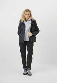 Vejrbestandig sort jakke over en grå fleece med lynlås, kombineret med sorte bukser og grå vandresko, sat mod en ensfarvet baggrund.