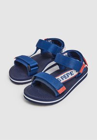 Modré látkové sandály se dvěma nastavitelnými pásky, texturovanou podrážkou a kontrastními červenými doplňky. Na páscích je logo.