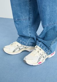 Baltas sporta apavi ar pelēkiem akcentiem un rozā detaļām, ar aizsargpārklājumu no acu materiāla un tekstūrētu zoli, apvienoti ar brīviem ziliem džinsu biksēm.